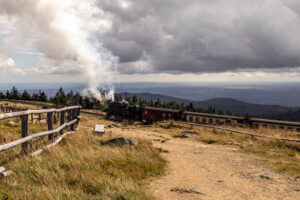 Historische Dampflok der Brockenbahn zieht Waggons um eine Kurve, wobei Rauch aufsteigt. Im Vordergrund sind ein sandiger Wanderweg und ein Holzzaun zu sehen, mit weitem Blick über den Harz im Hintergrund.