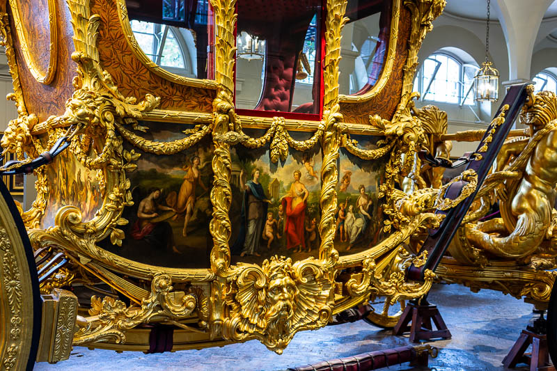 Nahaufnahme der kunstvoll bemalten Seitenpaneele der Gold State Coach, umrahmt von prunkvoll geschnitztem Blattgold. Zu sehen sind allegorische Szenen des italienischen Malers Cipriani.