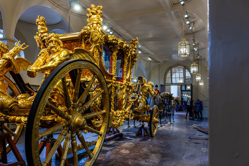 Perspektivische Ansicht der Gold State Coach von hinten in den Royal Mews London. Im Fokus stehen die vergoldeten Hinterräder, die Tritonen-Skulpturen mit Dreizack und die prachtvolle Verzierung des Gehäuses.