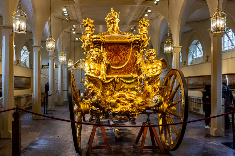 Rückansicht der Gold State Coach in den Royal Mews London. Die Aufnahme zeigt die großen Hinterräder, die vergoldeten Tritonen-Skulpturen mit Dreizack und das verzierte Rückpaneel der Kabine.