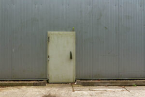 Kleine Tür in einer großen grauen Metallwand, mittig platziert, davor Betonboden